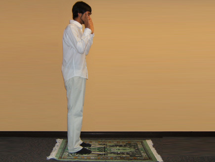 A man stands with his hands raised to his ears on a prayer mat